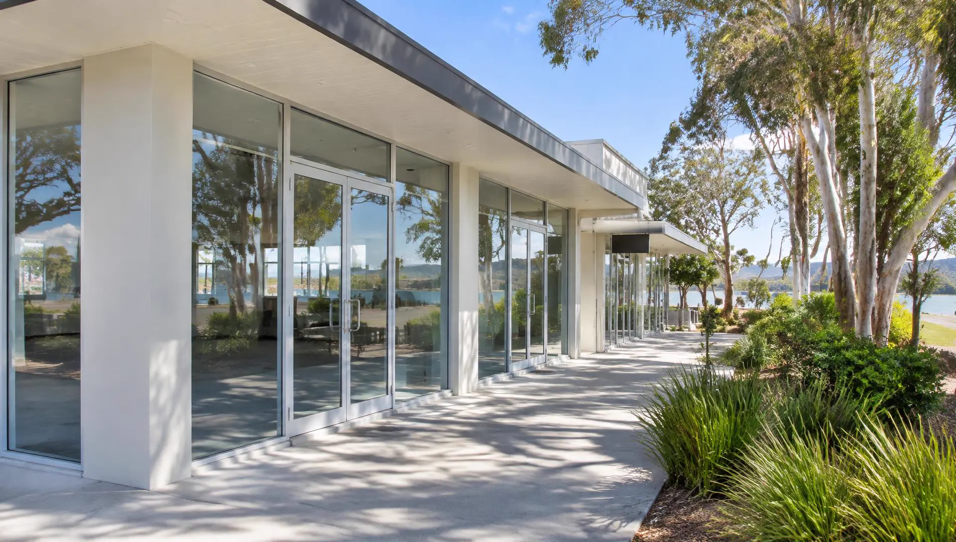Low-rise Central Coast shopfront with full-height glass and aluminium-framed doors beside eucalyptus trees and a waterway